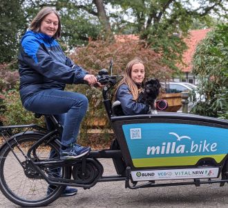 Eine Frau fährt ein milla-bike-Lastenrad mit einem Mädchen und einem Hund im Lastenkorb.