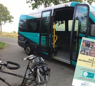 Ein Kleinbus mit geöffneten Türen lädt zum Einsteigen ein, davor steht ein Fahrrad und ein Plakat.