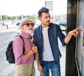 Junger Mann und blinder Senior mit weißem Rohr an Bushaltestelle in der Stadt.